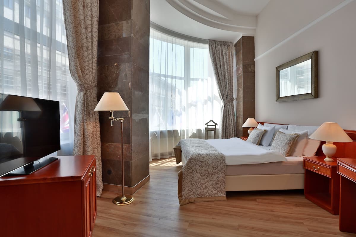 Habitación de hotel con cama, lámparas de noche, televisor y un ventanal con cortinas.