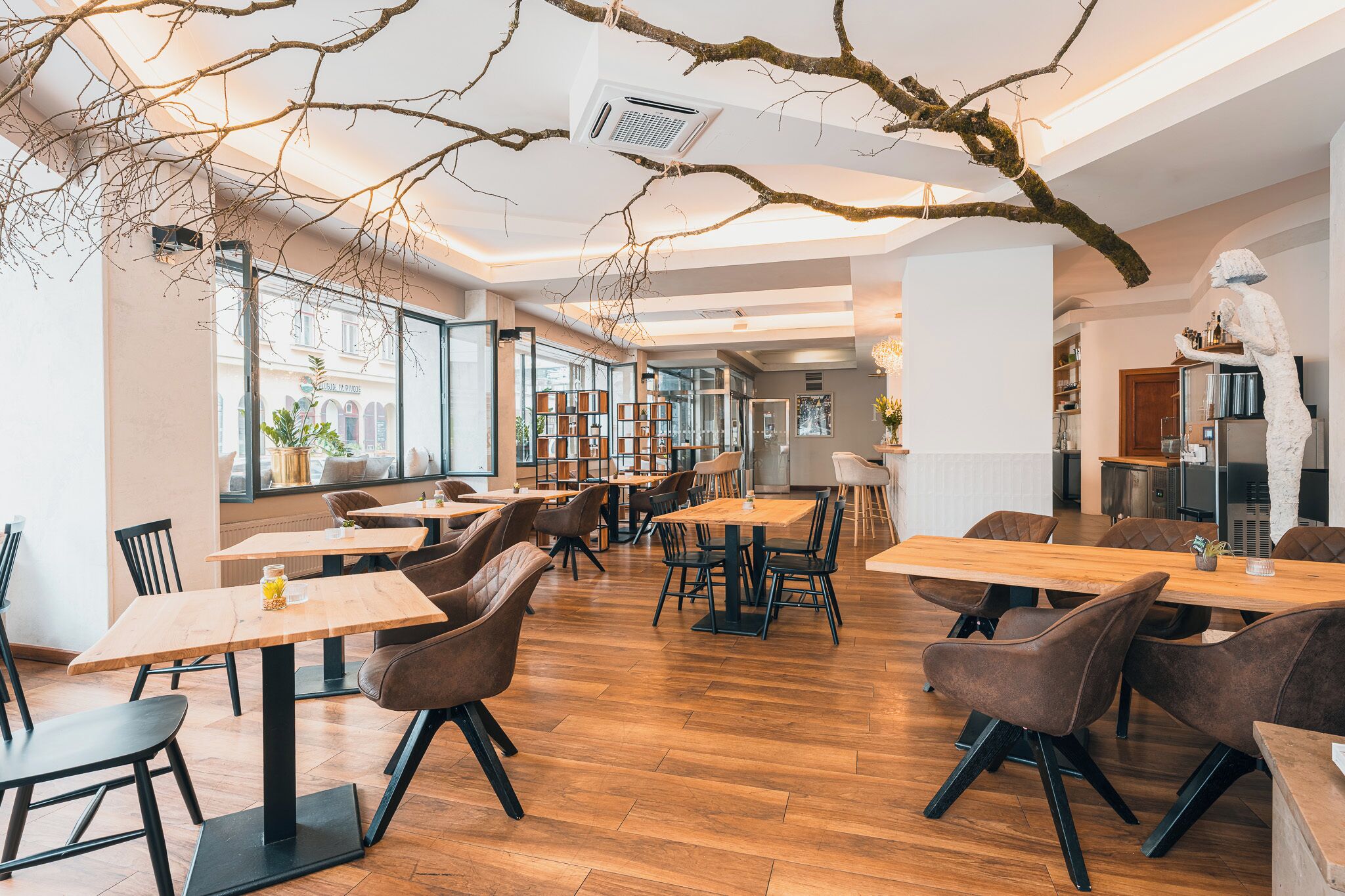 A modern restaurant interior with wooden tables and chairs, large windows, and a central bar area.