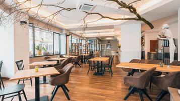 A modern restaurant interior with wooden tables and chairs, large windows, and a central bar area.