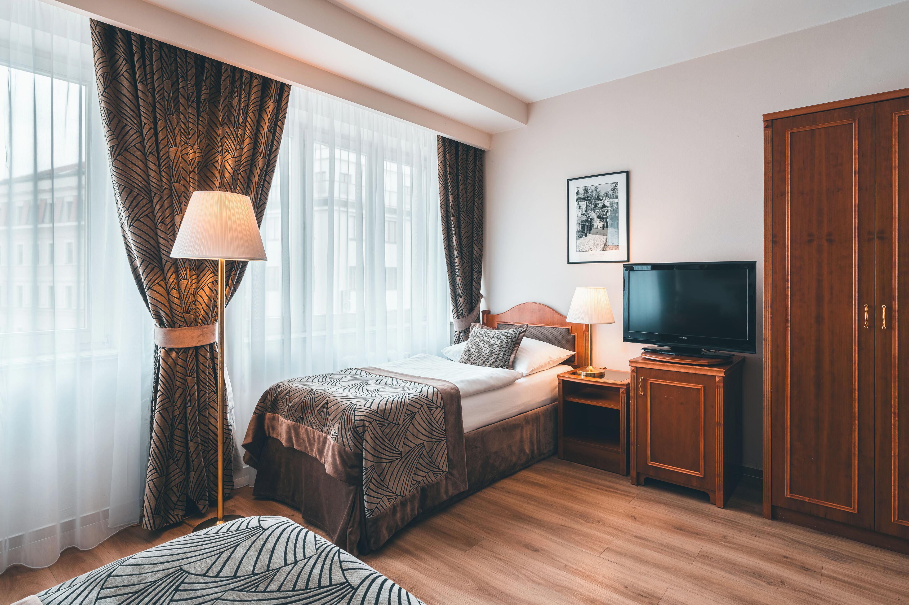 A hotel room with a bed, a television, a nightstand, a wardrobe, and a large window with curtains.