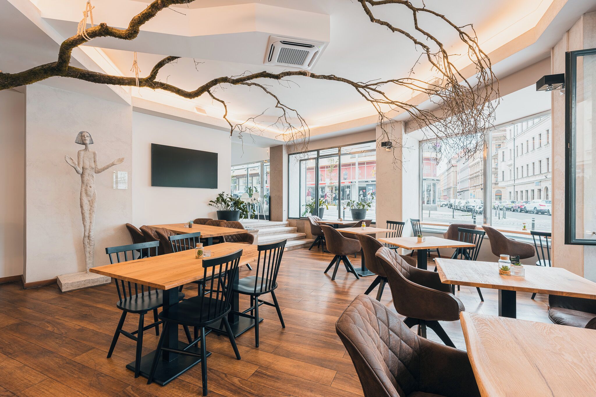 A modern cafe interior with wooden tables and chairs, large windows, and a hanging branch decoration.