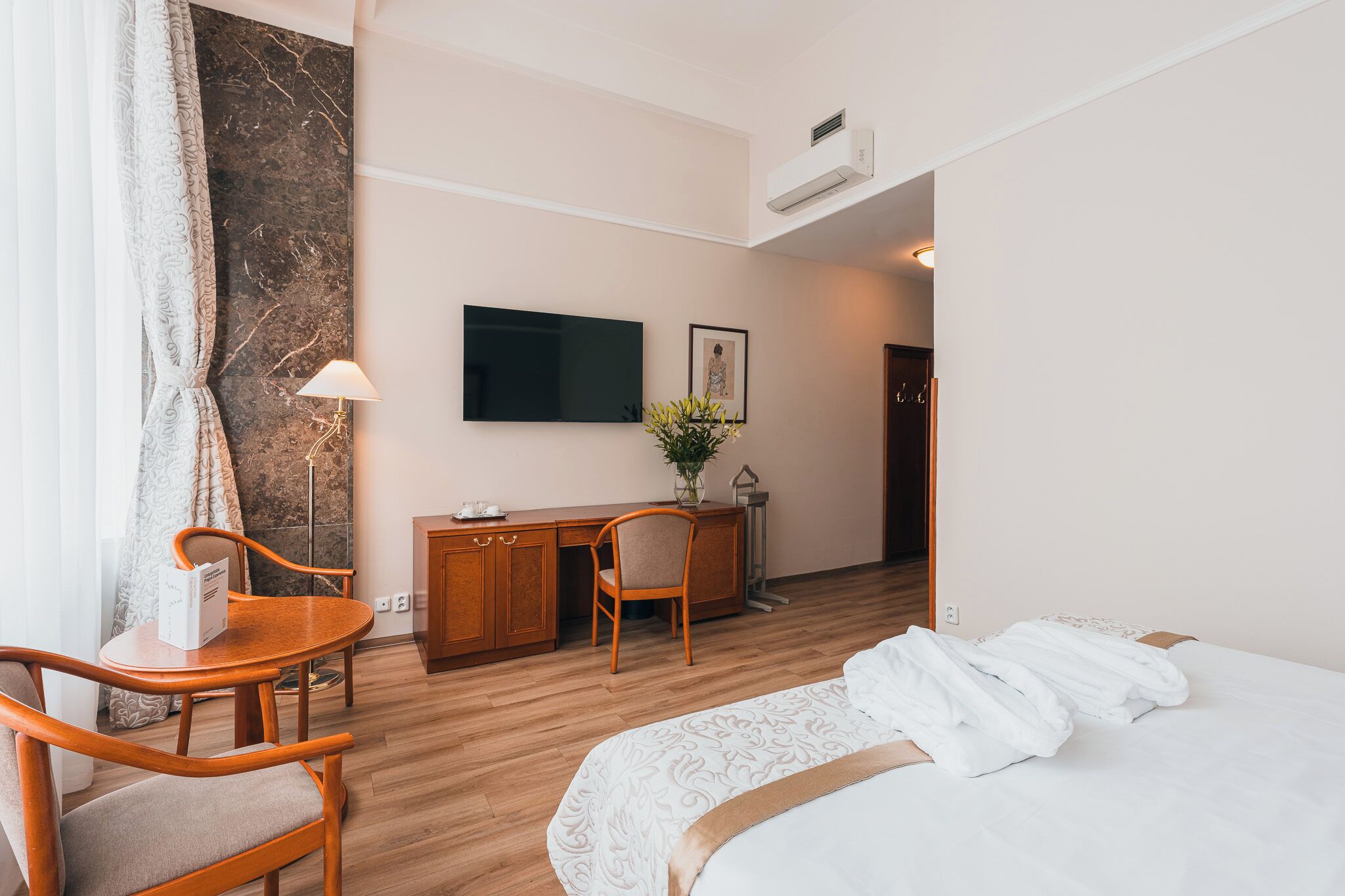 Ett hotellrum med en säng, ett skrivbord, en stol, en TV och en väggmonterad luftkonditioneringsanläggning.