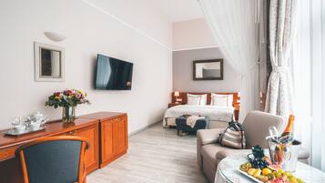 Quarto de hotel com uma cama, uma escrivaninha com cadeira, televisão, mesa de cabeceira com abajur, um vaso com flores e um prato de frutas sobre uma mesa.