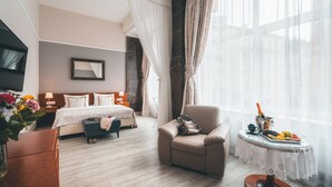 A hotel room with a bed, a sofa, a small table with a drink and fruit, and a view of buildings through large windows. - Hotel Belvedere (Prague)