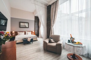 Ein Hotelzimmer mit Bett, einer Couch, einem kleinen Tisch mit Getränken und Früchten sowie Blick auf Gebäude durch große Fenster.