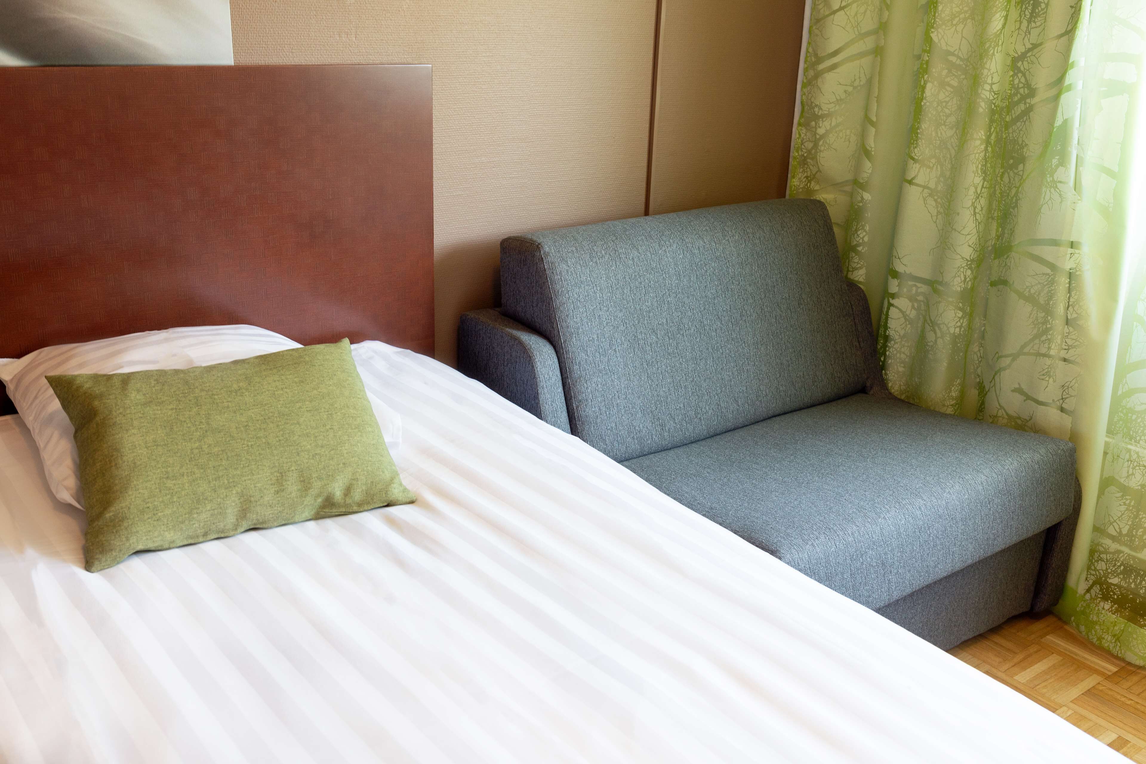 A bed with white bedding and a green pillow, a grey sofa, and a wooden floor.