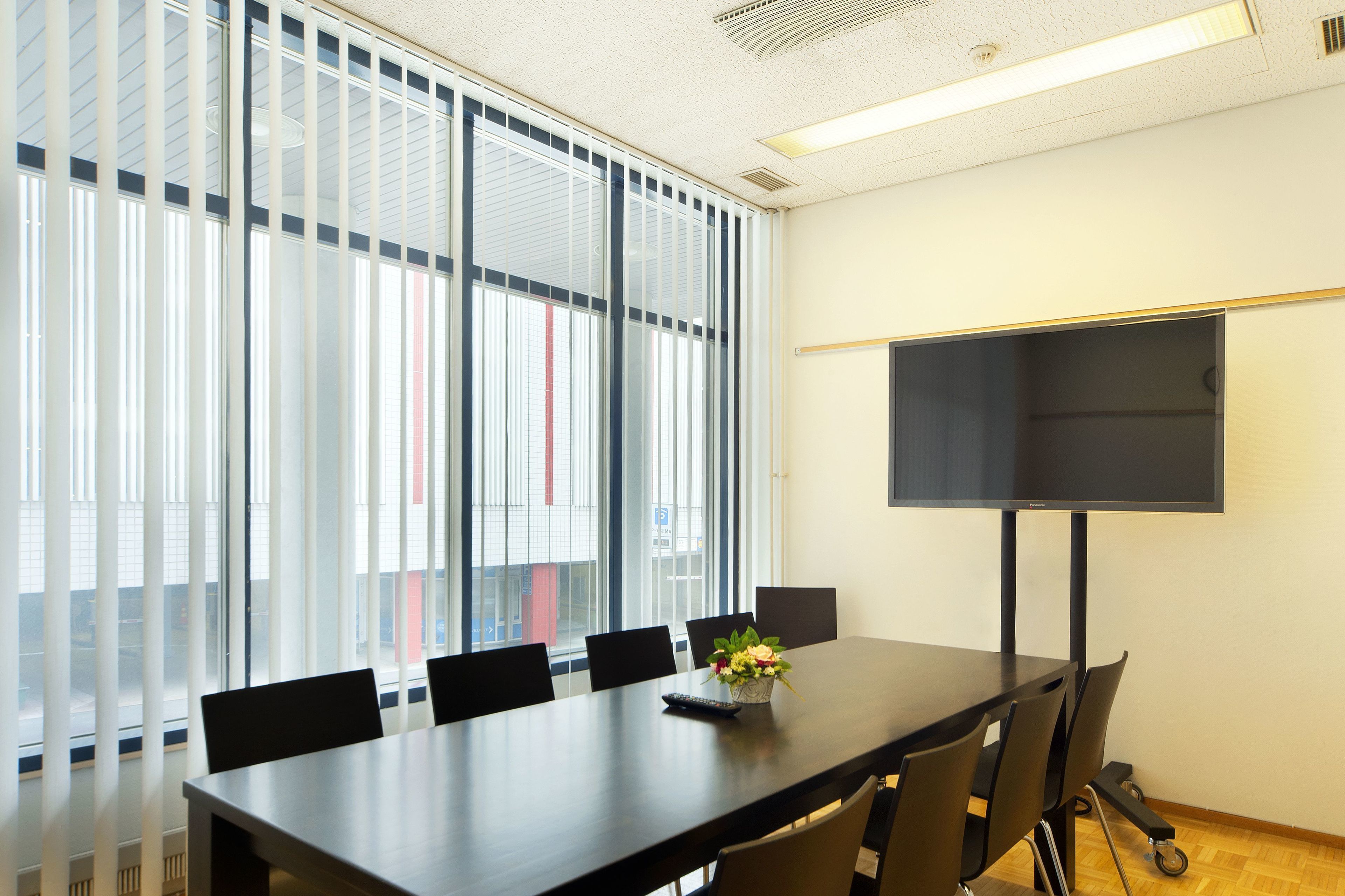 A conference room with a long table, six chairs, a flower vase, a remote control, and a large screen on a stand.