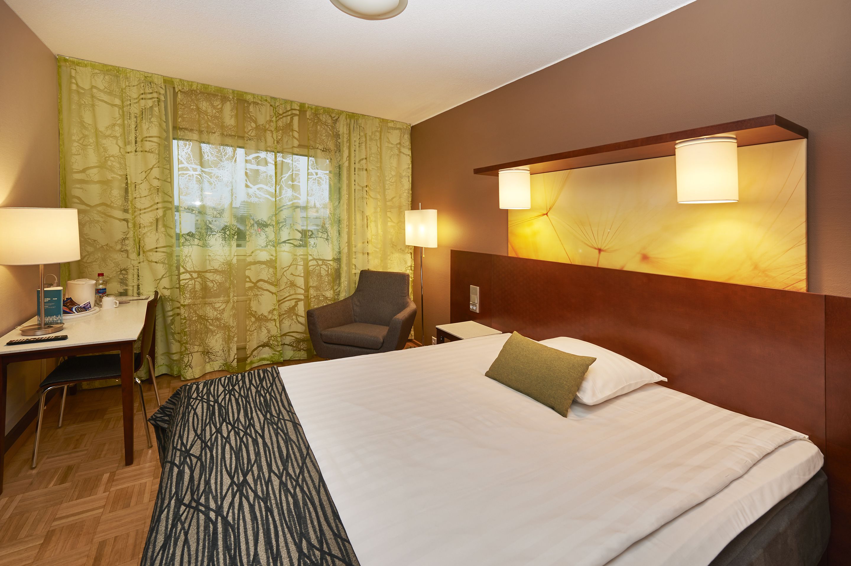Ein Hotelzimmer mit einem großen Bett, einem Schreibtisch mit Stuhl, einer Lampe und einem Fenster mit Vorhängen.