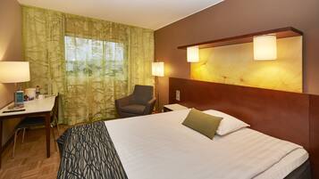 A hotel room with a large bed, a desk with a chair, a lamp, and a window with curtains.