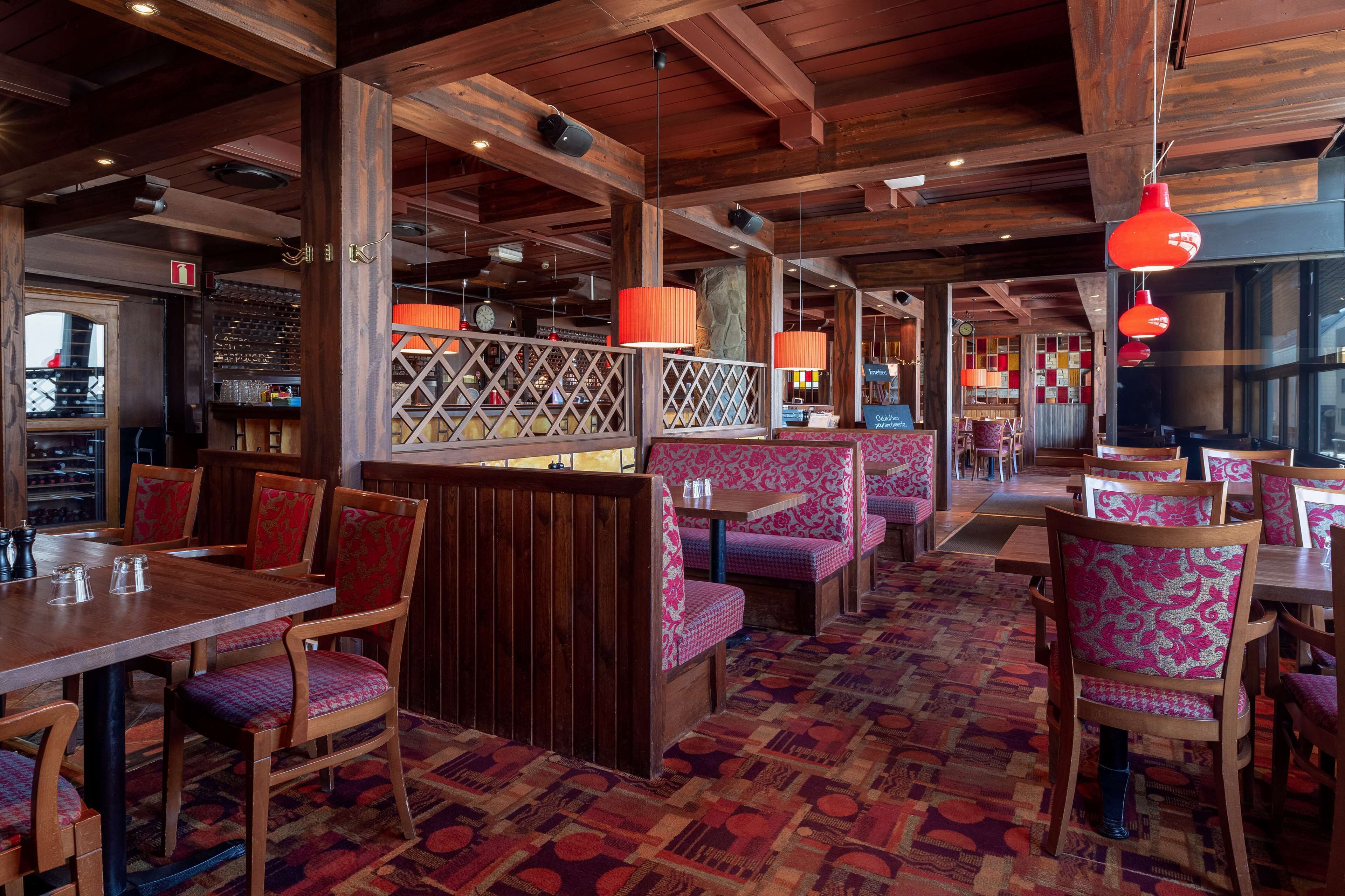 A restaurant interior with wooden tables and booths, patterned floor, and red pendant lighting.