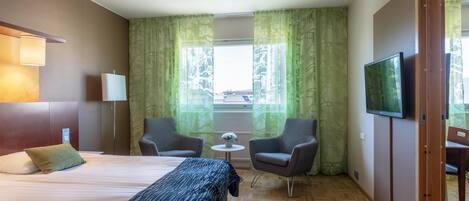 A hotel room with a bed, two armchairs, a small table with a plant, a TV, and a window with curtains.