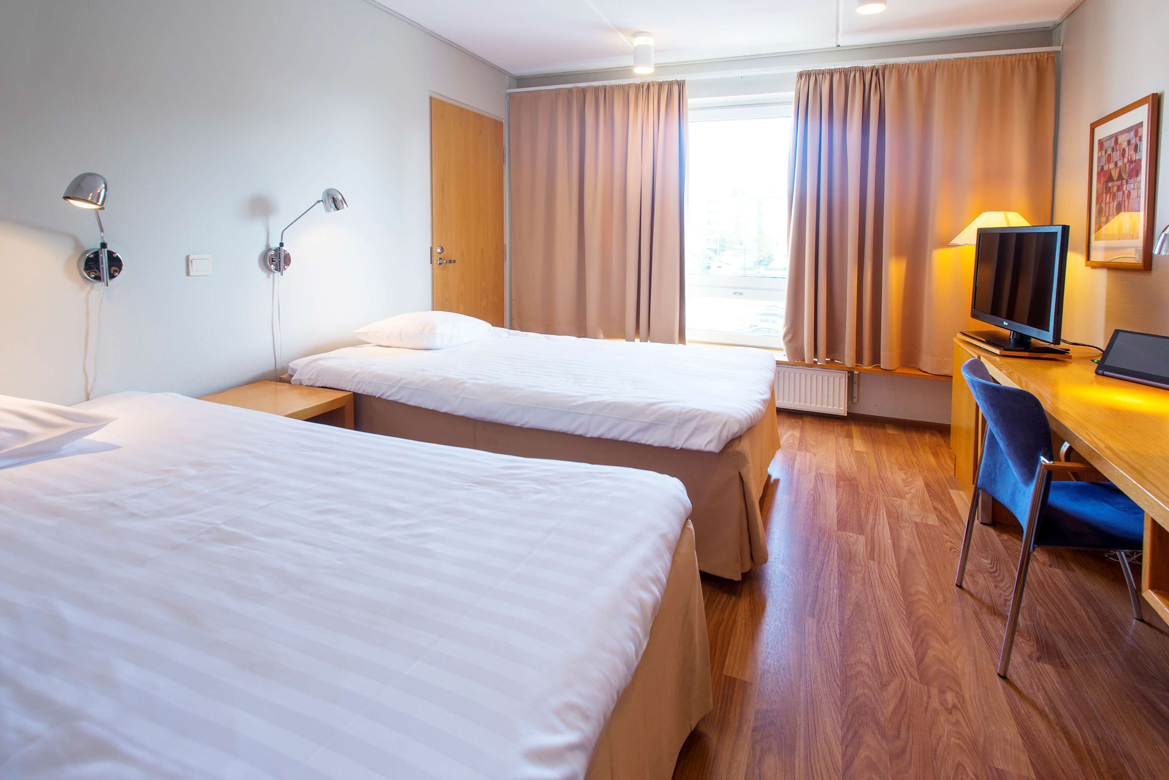 Ein Hotelzimmer mit zwei Betten, einem Schreibtisch mit Computer, einem Fernseher und zwei Wandlampen.