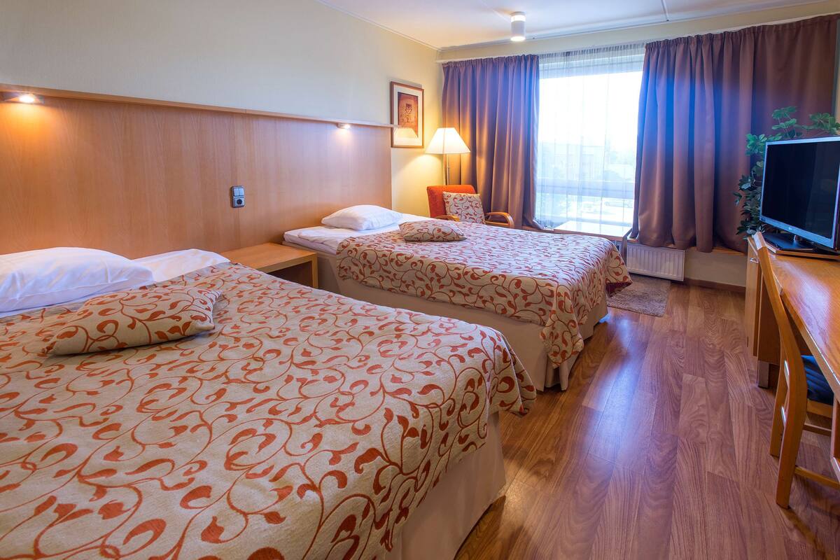 A hotel room with two beds, a TV, a lamp, and a window with curtains.