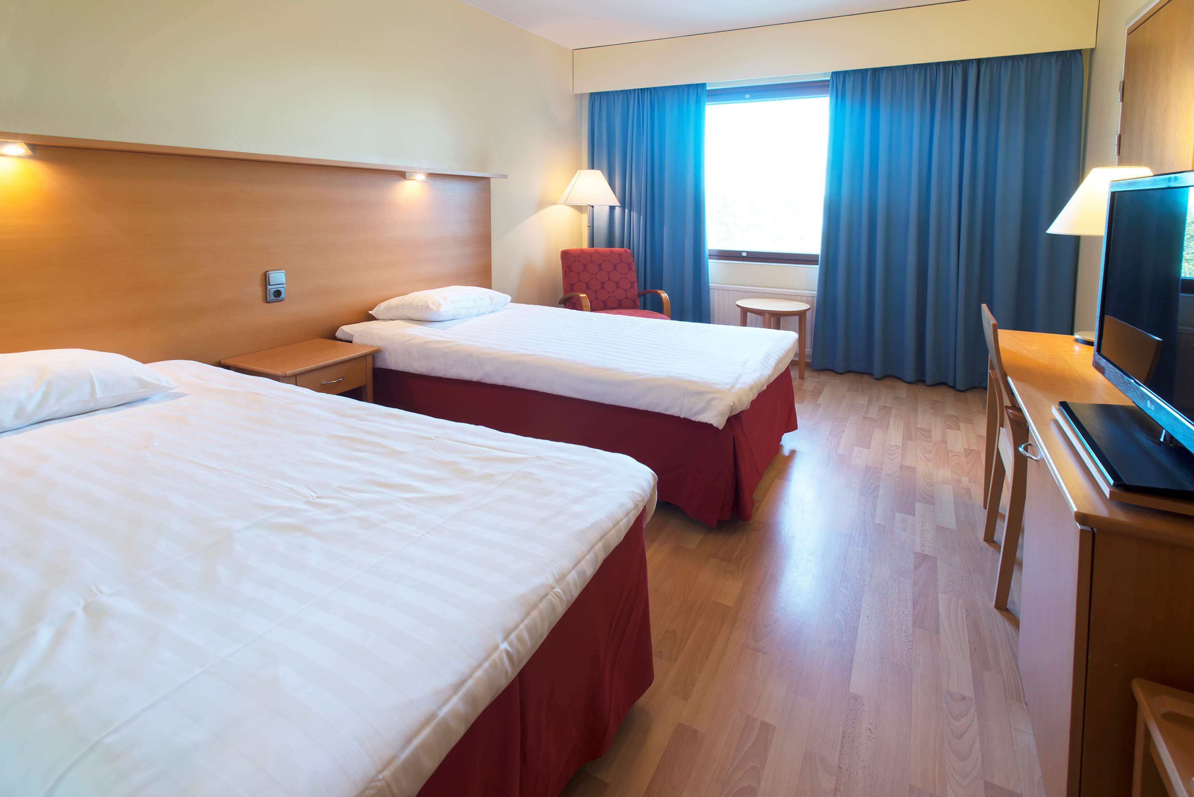 A hotel room with two beds, a desk, a chair, a TV, and a window with curtains.