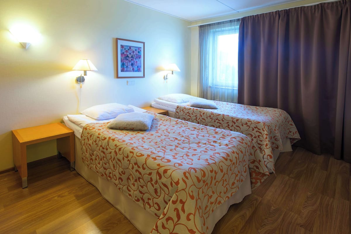 A hotel room with two beds, a nightstand, a lamp, a painting on the wall, and a window with curtains.