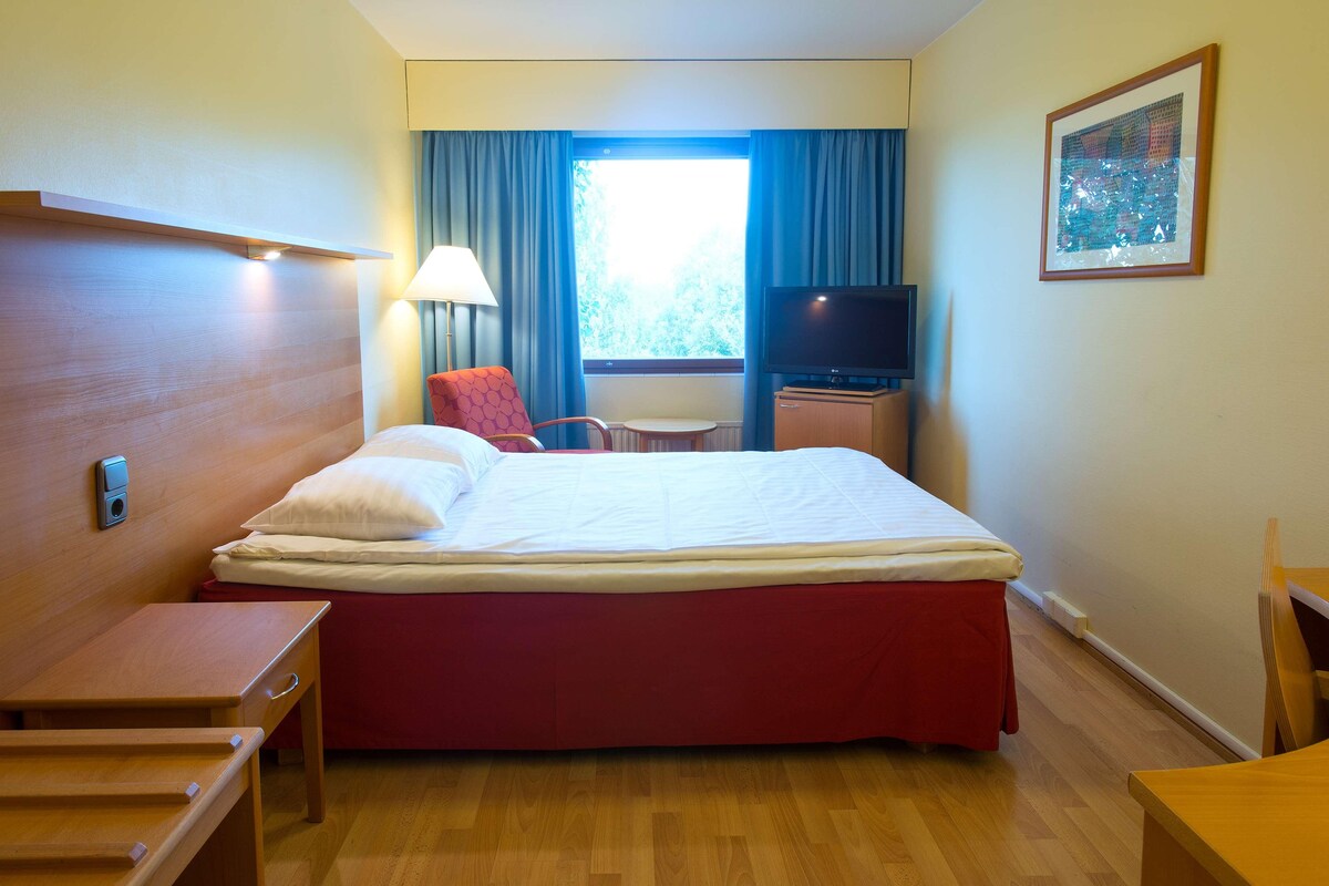 Quarto de hotel com cama, escrivaninha, cadeira, televisão e janela com cortinas.