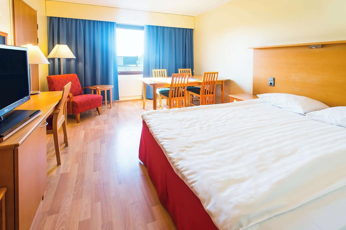 A hotel room with a bed, a desk with a TV, a chair, a table, and a window with curtains.