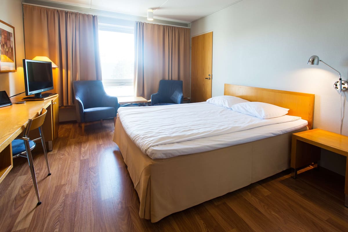 A hotel room with a bed, two armchairs, a desk with a computer, a TV, and a lamp.