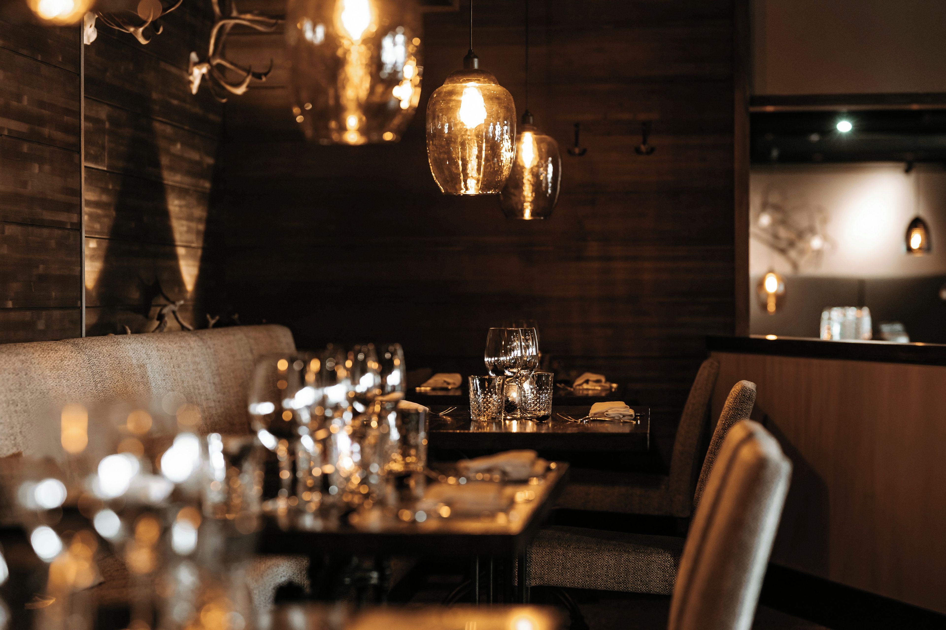 Un restaurante tenuemente iluminado con paredes de madera, lámparas colgantes y una larga mesa dispuesta con vasos y cubiertos.
