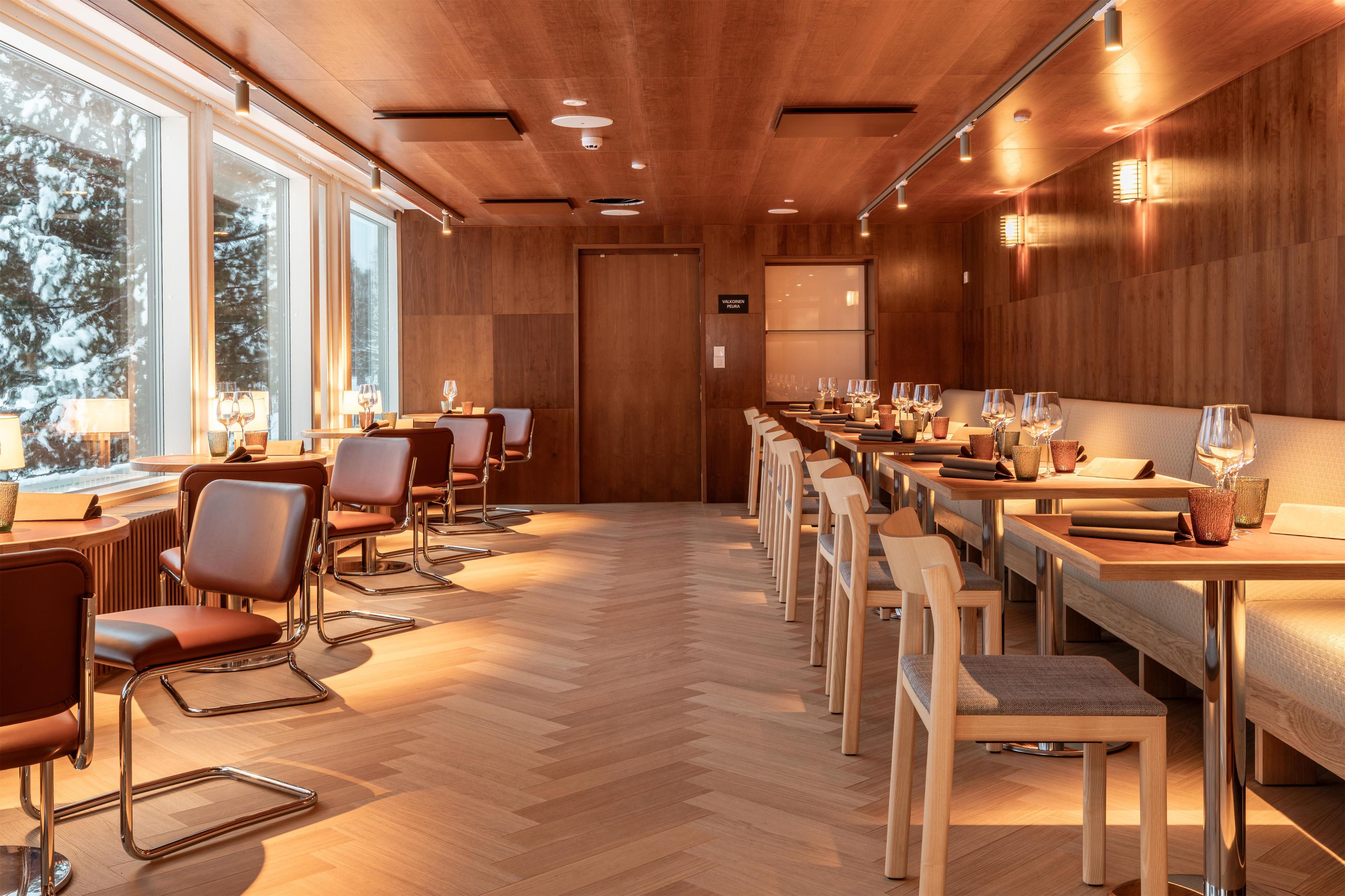 A modern restaurant with wooden paneling, large windows, and a long dining table with chairs.
