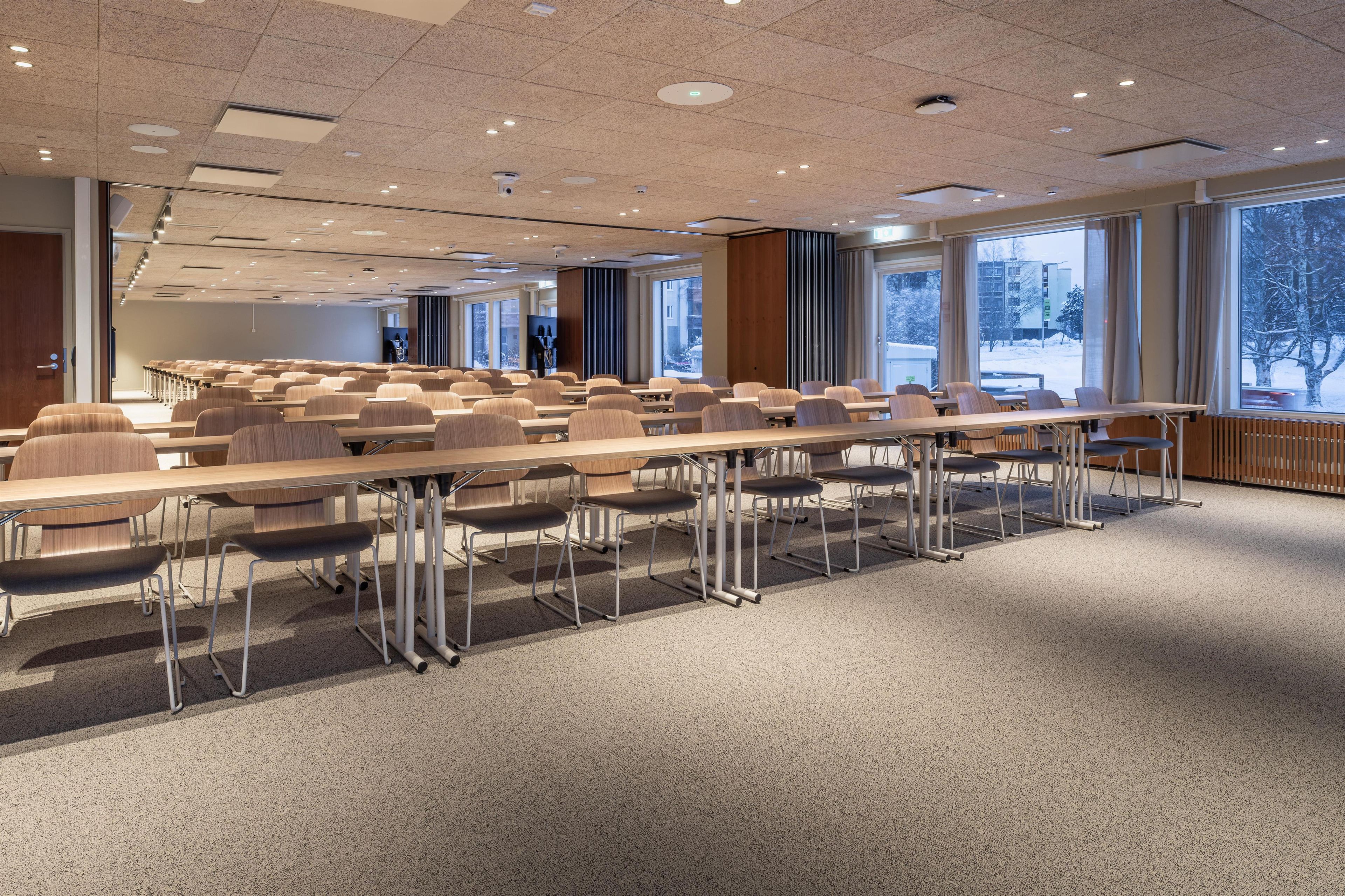 A conference room with rows of tables and chairs, large windows with a view of trees and buildings, and a ceiling with recessed lighting.