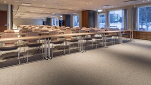 A conference room with rows of tables and chairs, large windows with a view of trees and buildings, and a ceiling with recessed lighting. - Scandic Pohjanhovi (Rovaniemi)