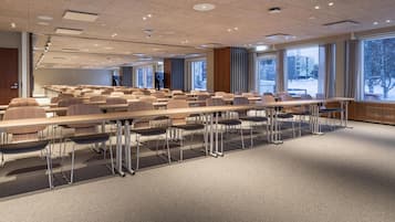 A conference room with rows of tables and chairs, large windows with a view of trees and buildings, and a ceiling with recessed lighting.