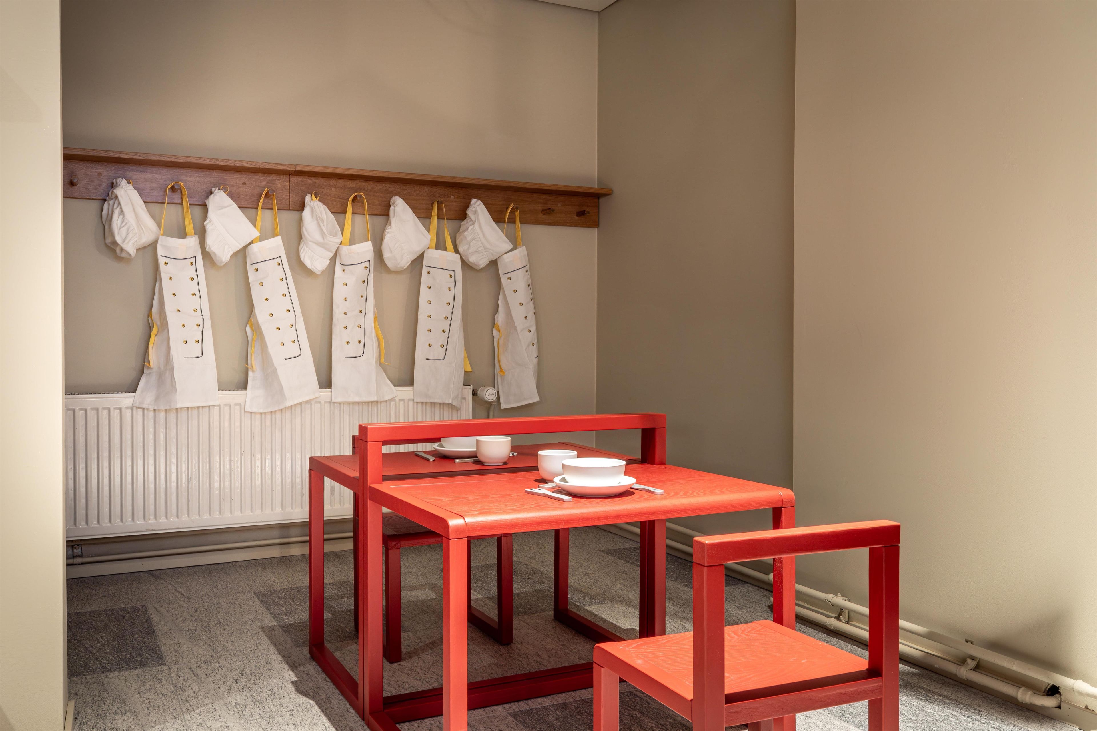 A room with a red table and chairs, a white radiator, and a row of white aprons hanging on a wooden rack.