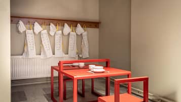 A room with a red table and chairs, a white radiator, and a row of white aprons hanging on a wooden rack.