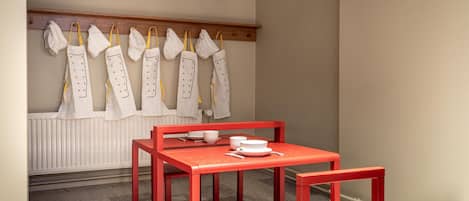 Una habitación con una mesa y sillas rojas, un radiador blanco y una fila de mandiles blancos colgados en un perchero de madera.
