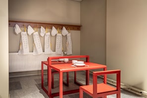 A room with a red table and chairs, a white radiator, and a row of white aprons hanging on a wooden rack.