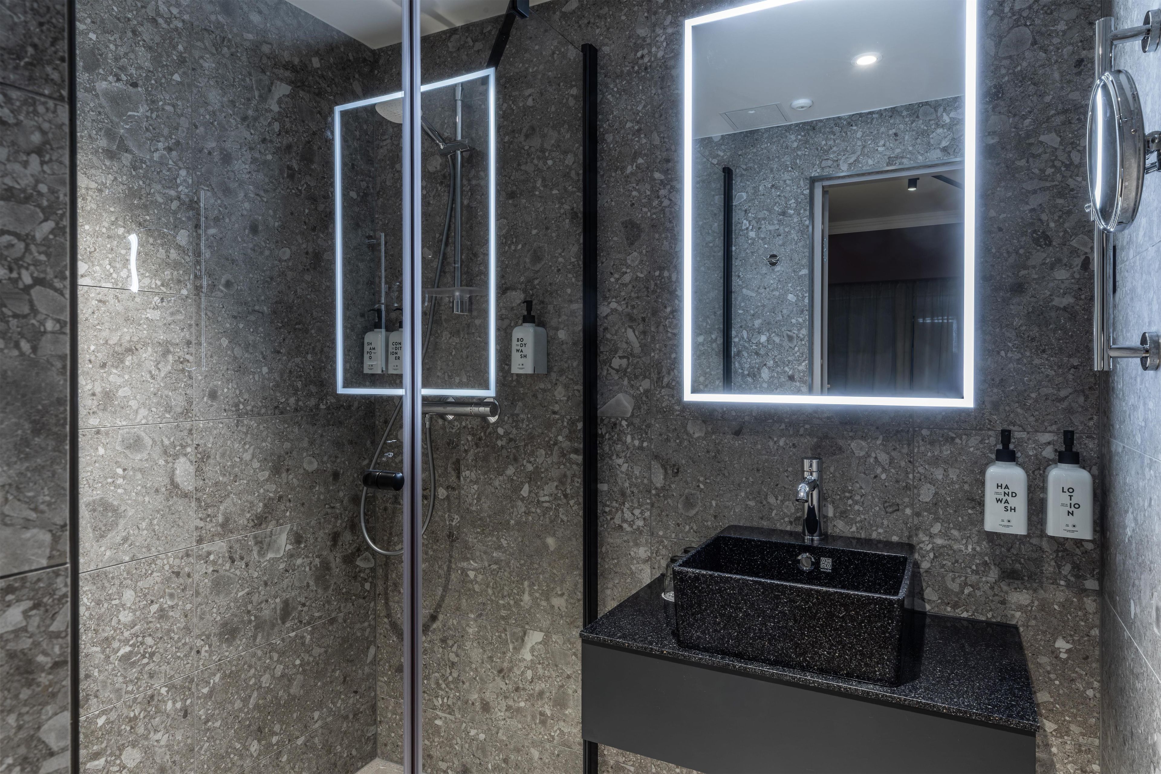 Un baño moderno con encimera de granito, ducha con cerramiento de vidrio y un espejo grande con iluminación integrada.