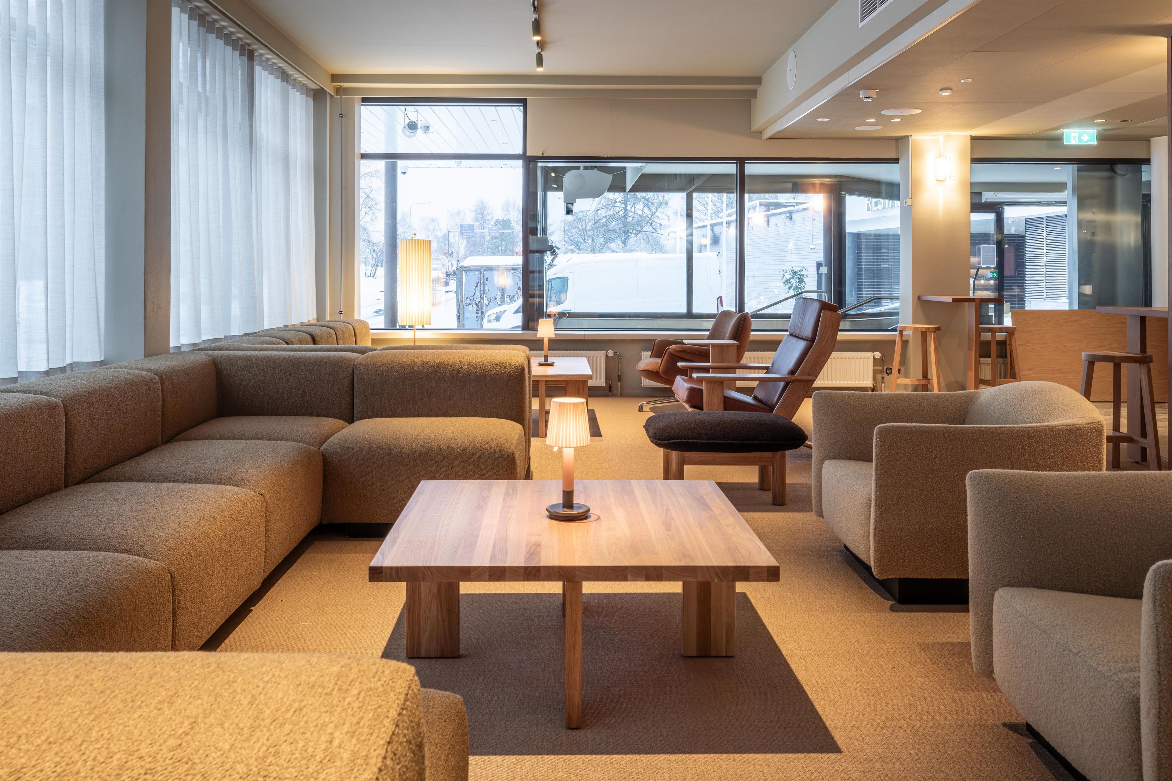 A modern hotel lobby with a large sectional sofa, a wooden coffee table, and several armchairs. Large windows provide a view of the outside.
