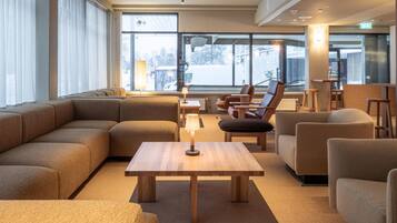 A modern hotel lobby with a large sectional sofa, a wooden coffee table, and several armchairs. Large windows provide a view of the outside.