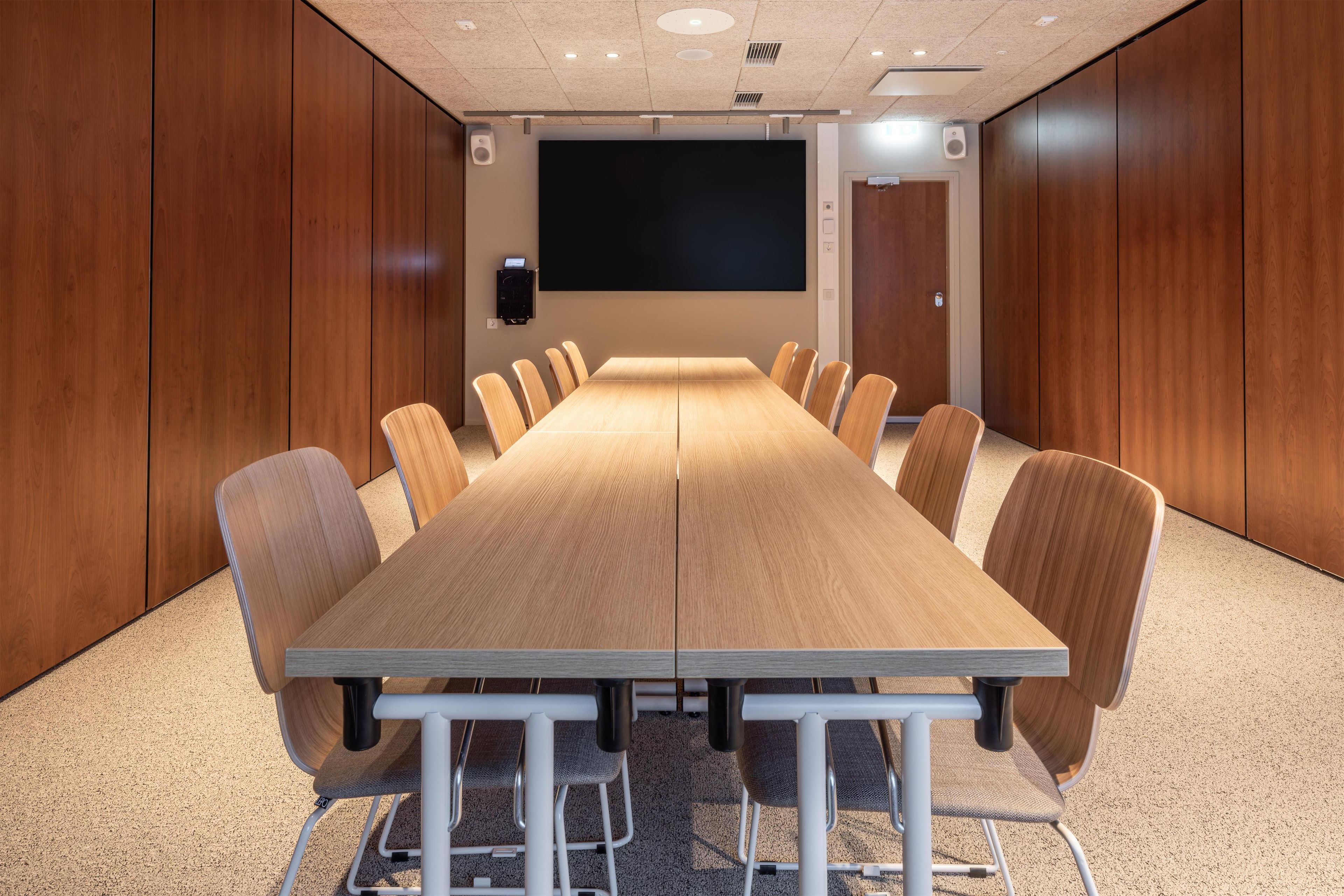 Ett konferensrum med ett långt bord, stolar, en projektorduk och högtalare.
