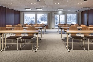 A conference room with multiple tables and chairs, large windows with a view of trees, and a carpeted floor.
