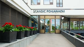 The exterior of a building with the name "SCANDIC POHJANHOVI" above the entrance. There are potted plants with red flowers in front of the entrance.