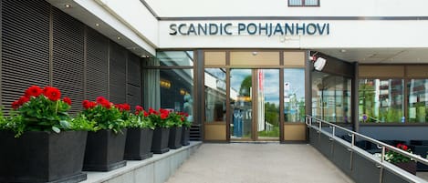 The exterior of a building with the name "SCANDIC POHJANHOVI" above the entrance. There are potted plants with red flowers in front of the entrance.