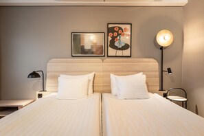 Habitación de hotel con dos camas, cabecero, mesita de noche, lámpara, reloj de pared y dos cuadros enmarcados.