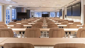 Un salón de conferencias con filas de mesas y sillas de madera, ventanas amplias y un televisor de pantalla plana.
