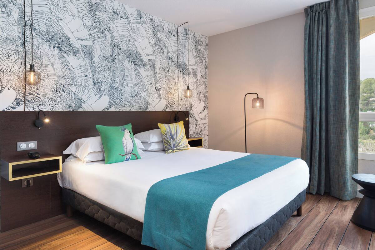 Une chambre d’hôtel moderne dotée d’un grand lit, d’un parquet en bois, d’un mur orné d’un motif de feuilles et offrant une vue sur l’extérieur.