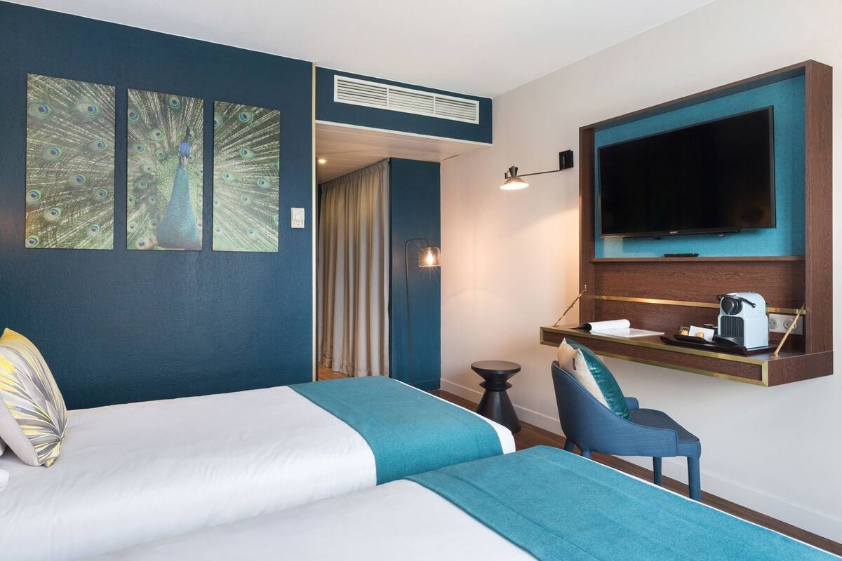 Une chambre d’hôtel avec un lit, deux tableaux représentant des paons, une télévision, un bureau avec une machine à café et une chaise.