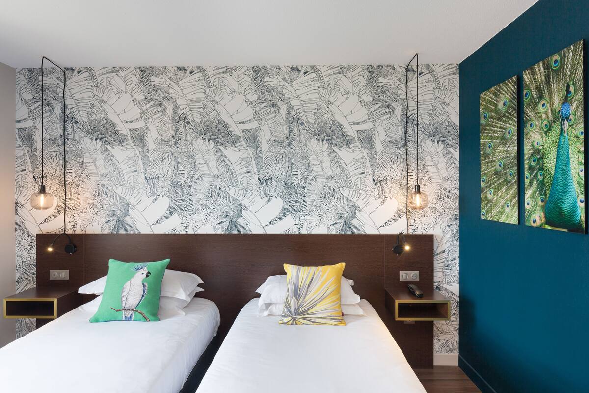 Une chambre d’hôtel avec deux lits, un tableau représentant un paon et un papier peint à motifs de feuilles.