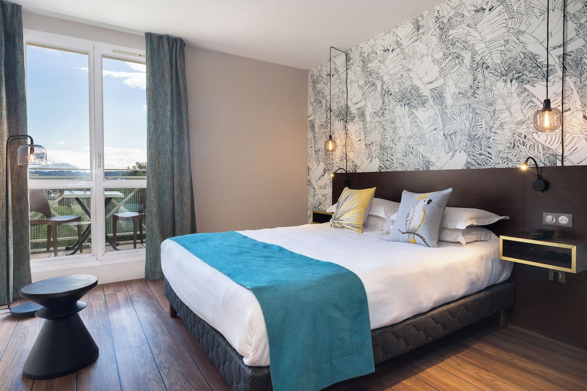 Une chambre d’hôtel avec un grand lit, une vue par la fenêtre, une petite table et une table de chevet avec une lampe.