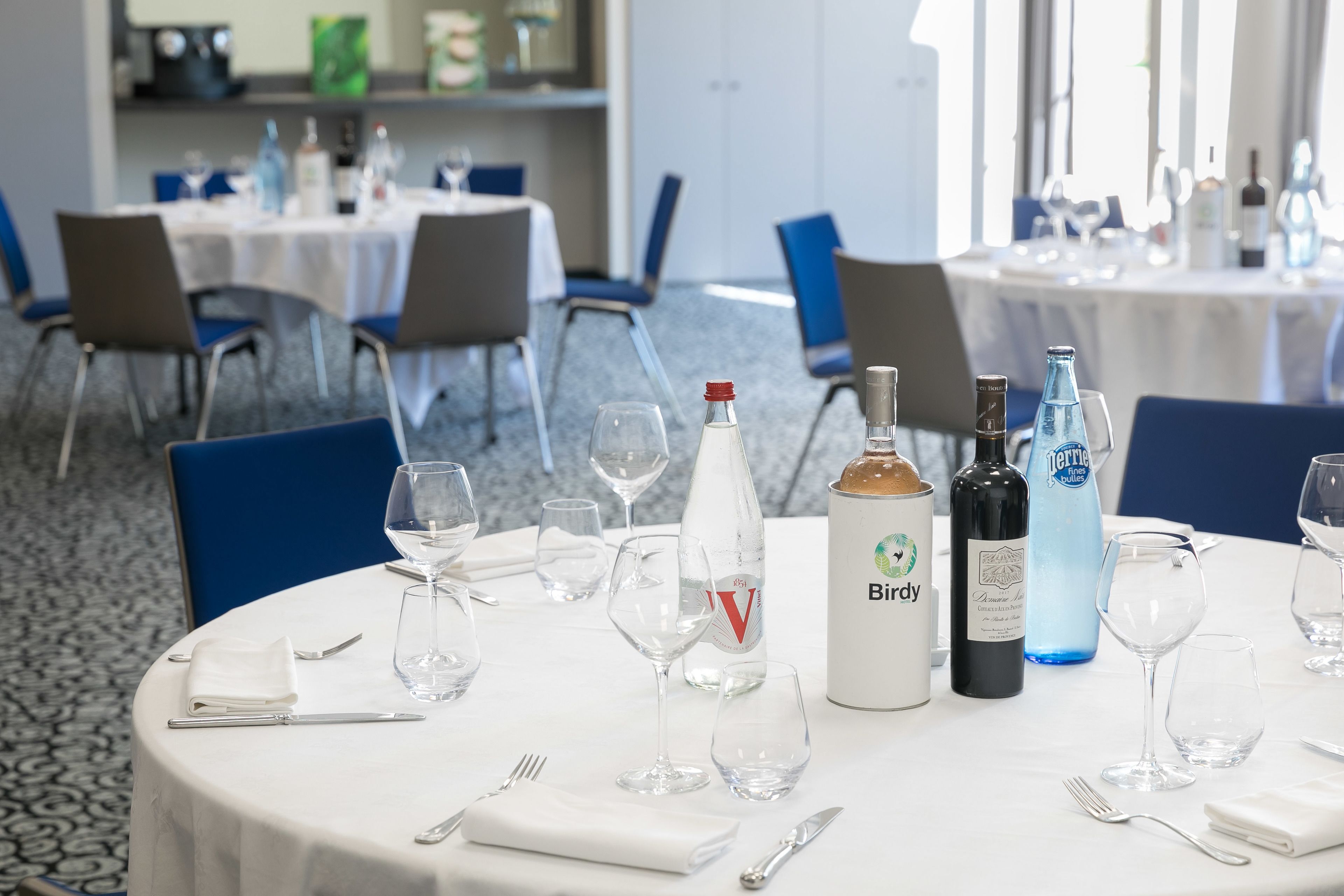 Ein Essbereich mit runden Tischen, gedeckt für eine Mahlzeit, jeder Tisch mit weißer Tischdecke, blaue Stühle und leere Weingläser.