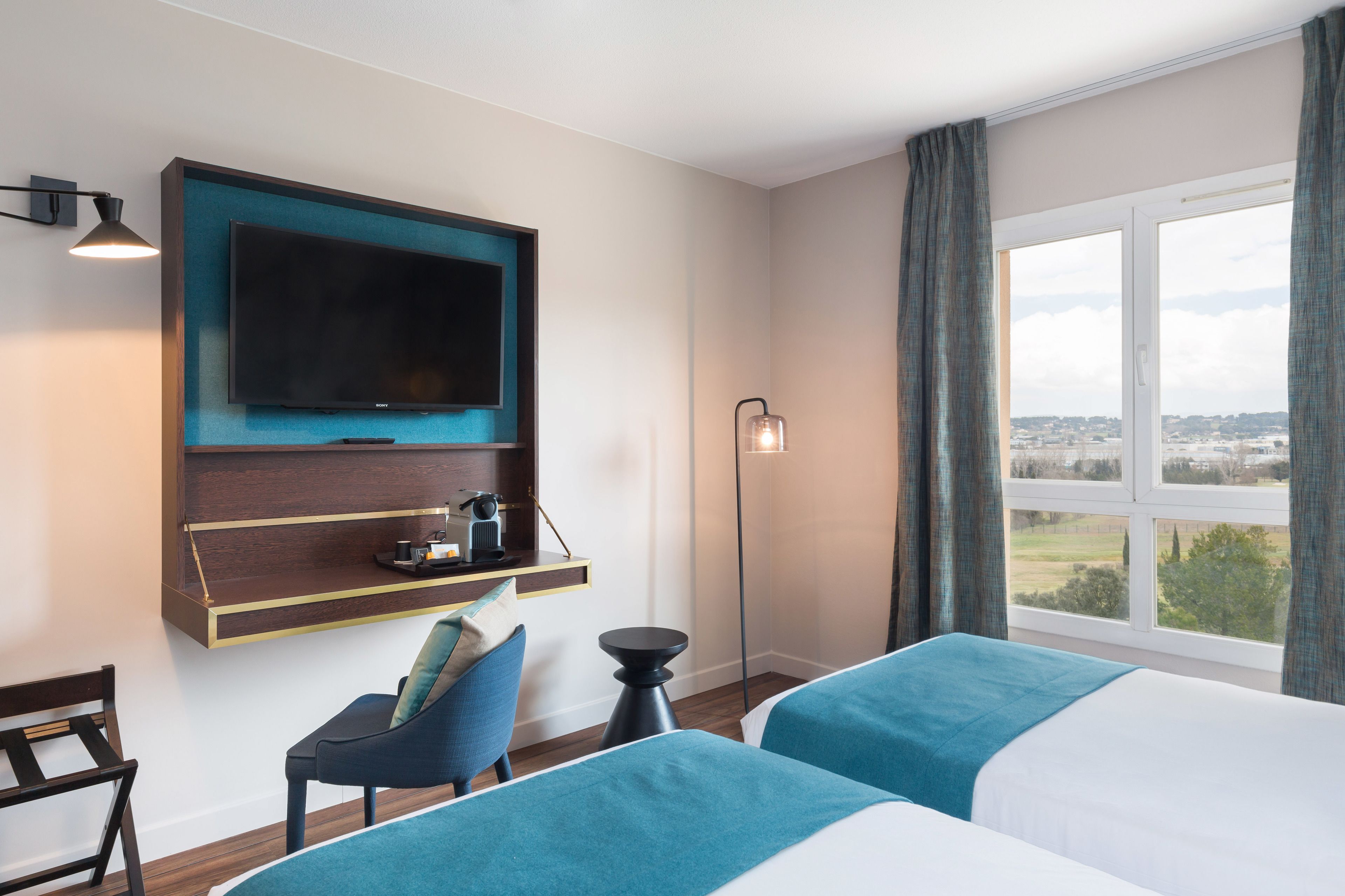 Une chambre d’hôtel avec un lit, une chaise, un bureau doté d’un téléviseur à écran plat, une lampe et une fenêtre donnant sur un paysage agréable.