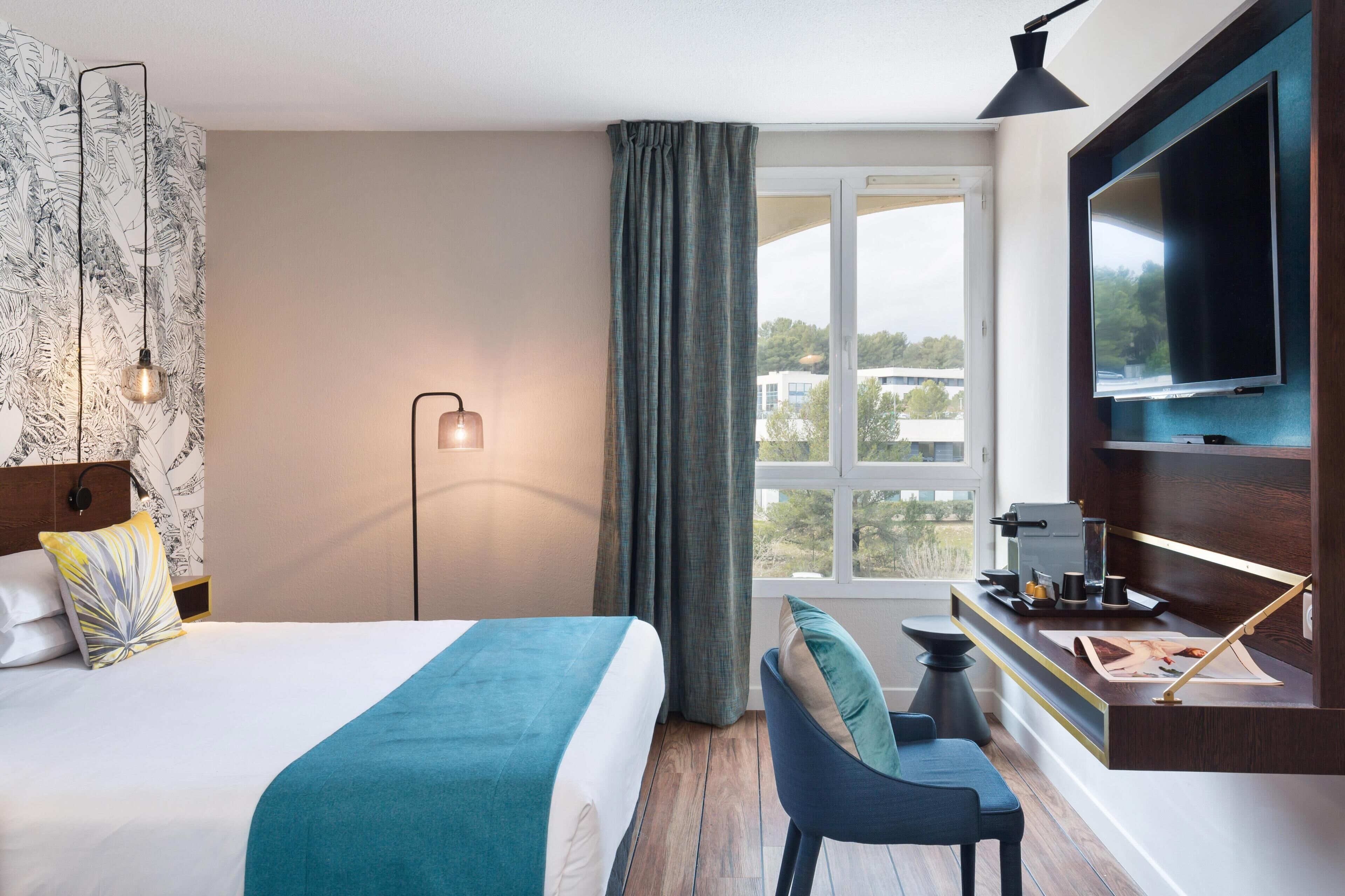 Une chambre d’hôtel comprenant un lit, un bureau, une chaise, une télévision, une fenêtre avec des rideaux et un mur orné d’un motif de feuilles.