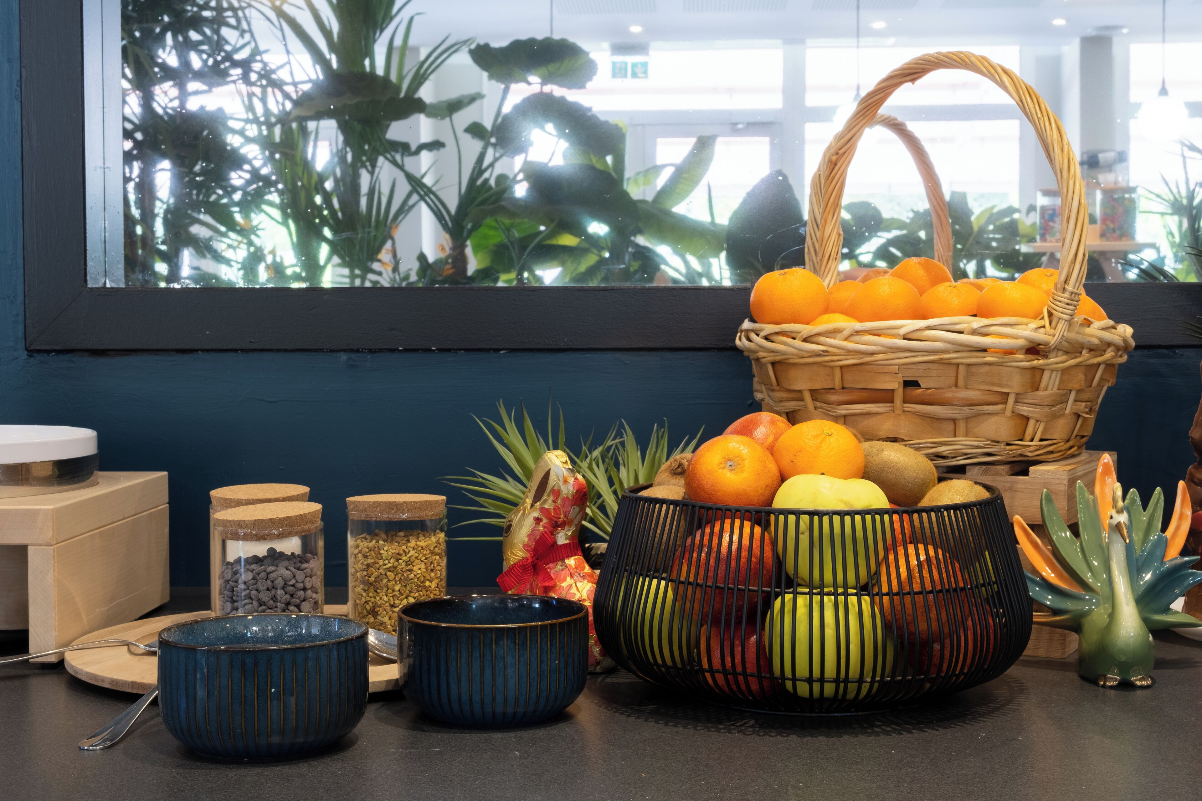 A kitchen counter with various bowls, a wicker basket with oranges, and a window with plants.
