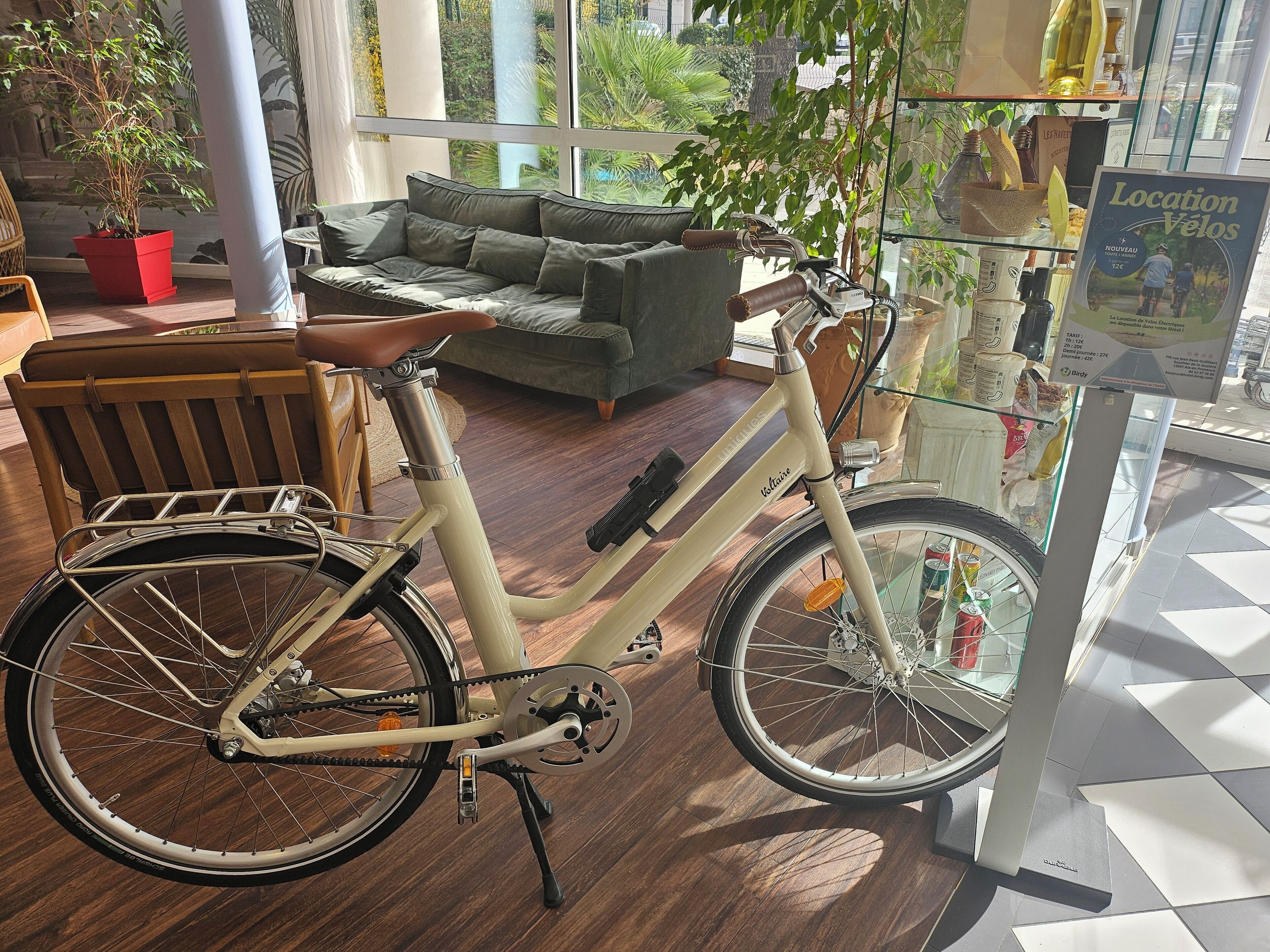En ældre cykel med kurv og sadel, placeret indendørs nær en glasmontre med skiltet "Location Vélos".
