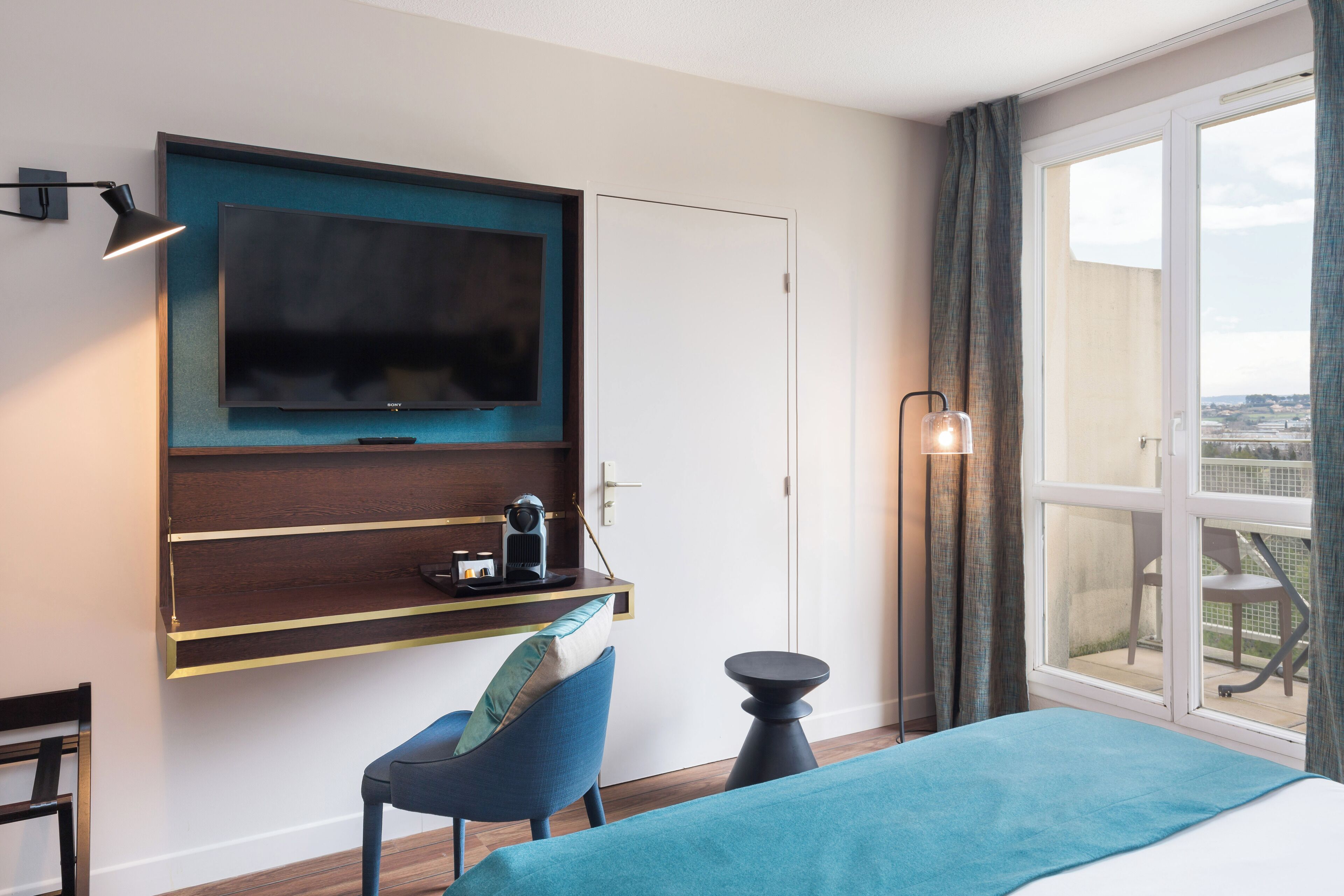 A modern hotel room with a large bed, a desk with a flat-screen TV, a chair, and a balcony with a view.